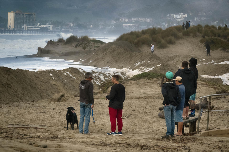 Residents stand on sand berms and watch as turbulent surf pounds the coast near the Ventura Pier, Saturday, Dec. 30, 2023, in Ventura, Calif. (AP Photo/Richard Vogel)