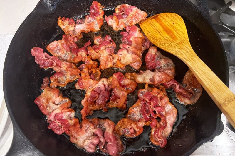 Bacon cooking in skillet with wooden spoon