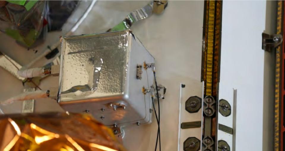 a small box wrapped in metallic foil on the side of a tall rectangular spacecraft on four legs