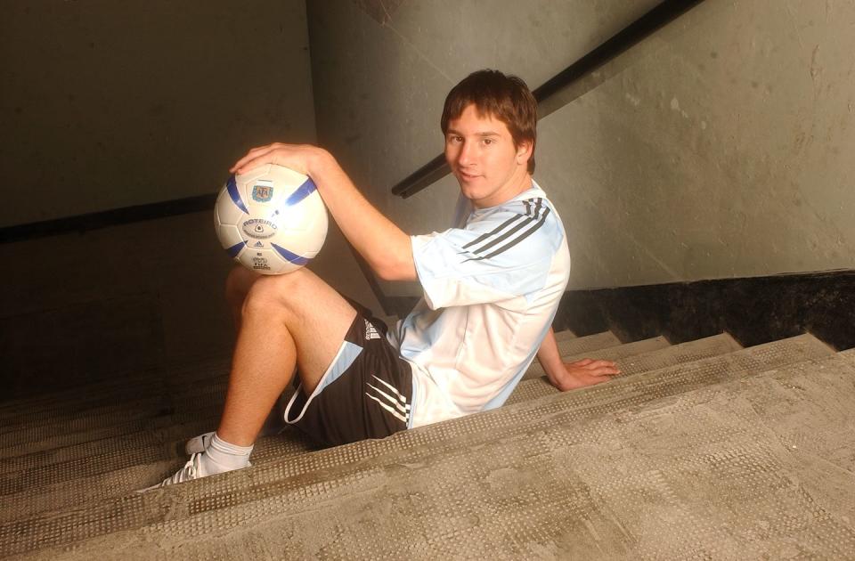 lionel messi sits in a stairwell and holds a soccer ball on his knees, he smiles at the camera and wears an argentina national team uniform