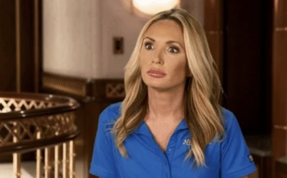 A woman with long blond hair wearing a blue polo shirt stands in an interior setting, looking surprised