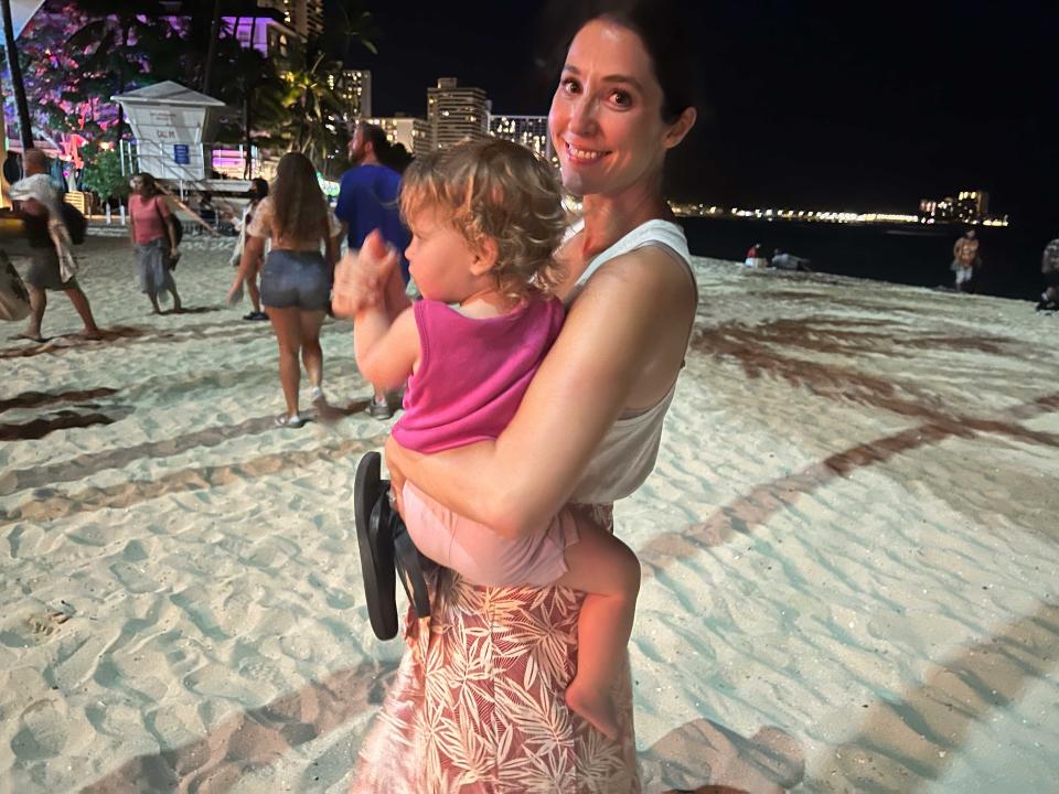 A woman smiling for a photo on the beach while holding her child.