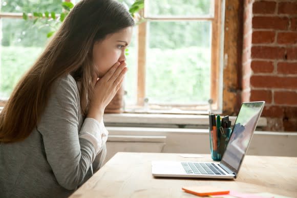 Woman looking at laptop and covering her mouth as if shocked