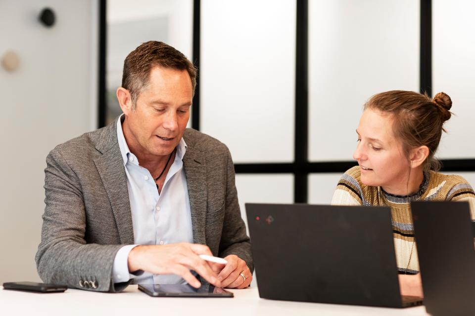 mark bula wearing a grey blazer and button down discussing something on a laptop with his colleague