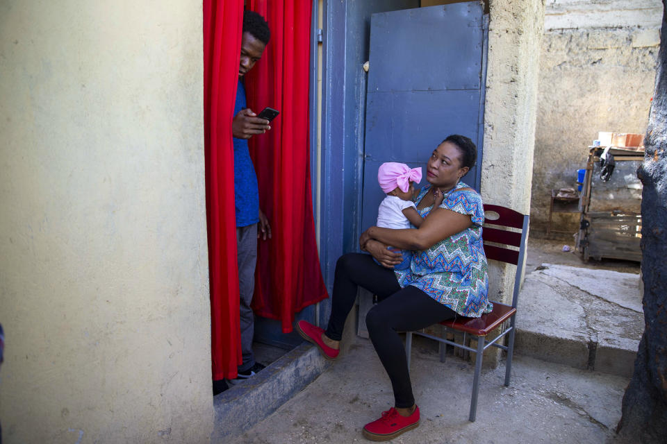Verty chats on his phone while his wife Saint Jean holds up her 1-year-old daughter at their house in Port-au-Prince, Haiti, Tuesday, Aug. 25, 2020. The Trump administration has sharply increased its use of hotels to detain immigrant children before expelling them from the United States during the coronavirus pandemic. Verty says government contractors at a hotel where he was detained gave his family, including his daughter, cups of ice to eat to pass temperature checks prior to their deportation flight, even though they had tested negative for COVID-19. (AP Photo/Dieu Nalio Chery)