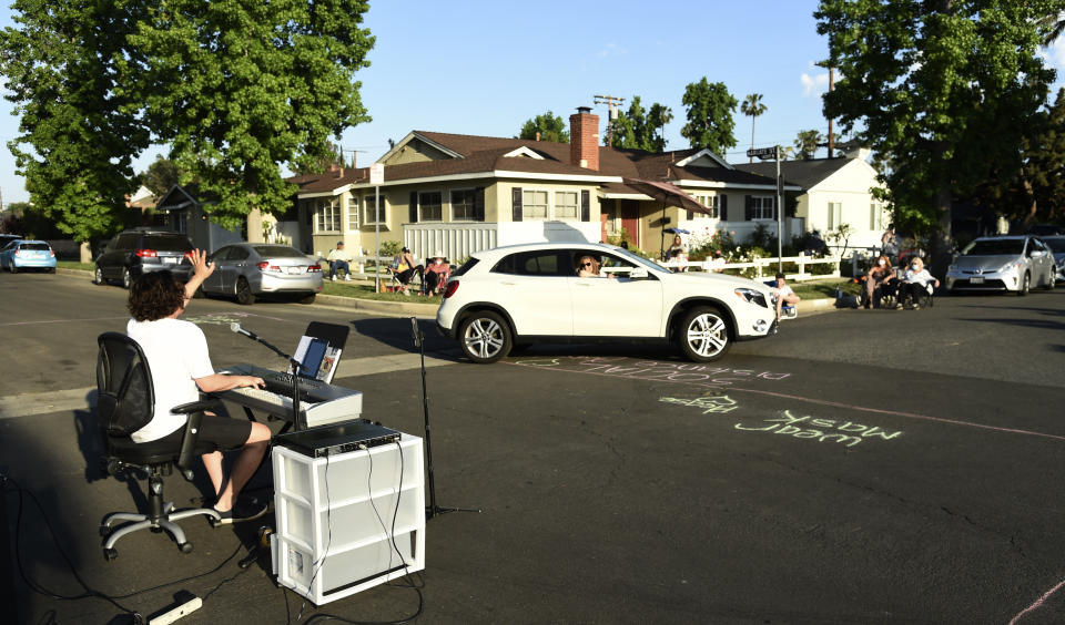 In this May 9, 2020 photo, musician Adam Chester, left, waves to a neighbor driving by as he performs his weekly neighborhood concert in the Sherman Oaks section of Los Angeles. Normally, Chester is a surrogate Elton John, who sings and plays the rock superstar's parts at rehearsals. With that work on hold, Chester has been giving concerts to his neighbors from a safe social distance in front of his house. (AP Photo/Chris Pizzello)