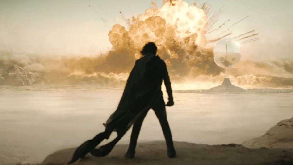  A man in cloak stands in the desert watching a massive explosion in the distance. 