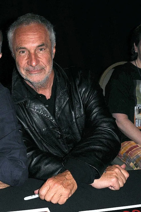 Two men, Steve Bisley and John Jarratt, sit at a table, smiling at the camera. Steve wears a vest and shirt, and John wears a leather jacket