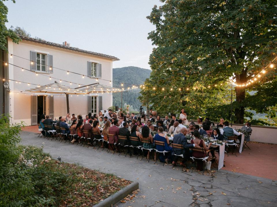 A large group of people sit at tables with strings of lights above them.