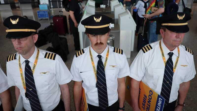 Three airline pilots in white shirts with black caps and black and white striped ties stand shoulder to shoulder in front of airport check in.