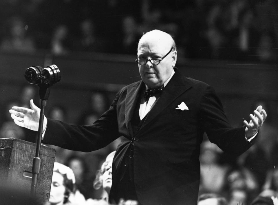 black and white photo of Churchill in suit with bow tie behind a microphone with both arms raised as if to say, "What?"