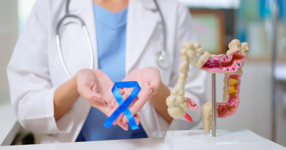 close up of female doctor hand wear white coat holds blue ribbon in front of her chest with colon model on table