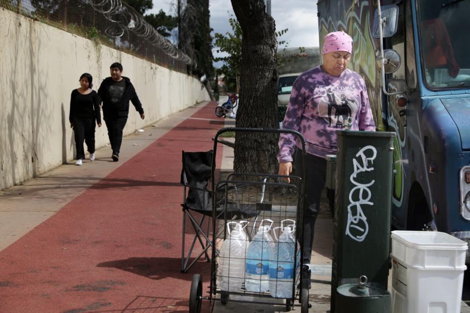 A woman with a cart of plastic jugs near a fountain on a sidewalk