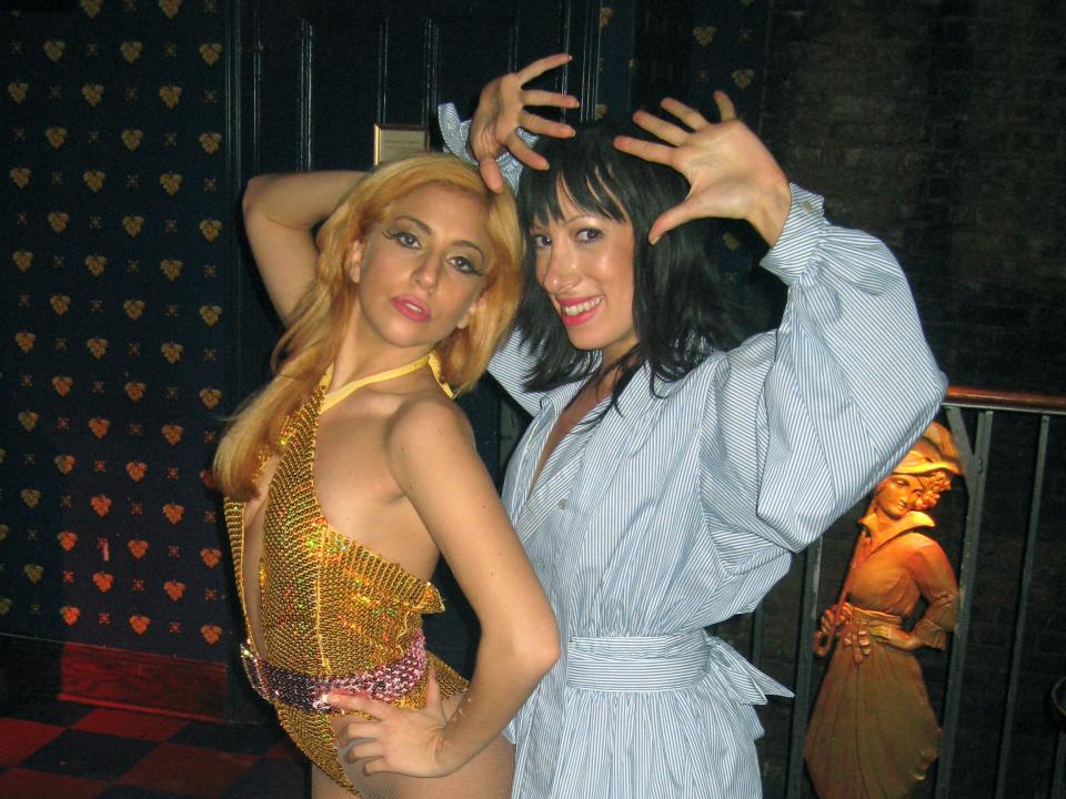 Gaga with yellow hair and a gold costume posing next to Lady Starlight in a blue dress with her hands up.