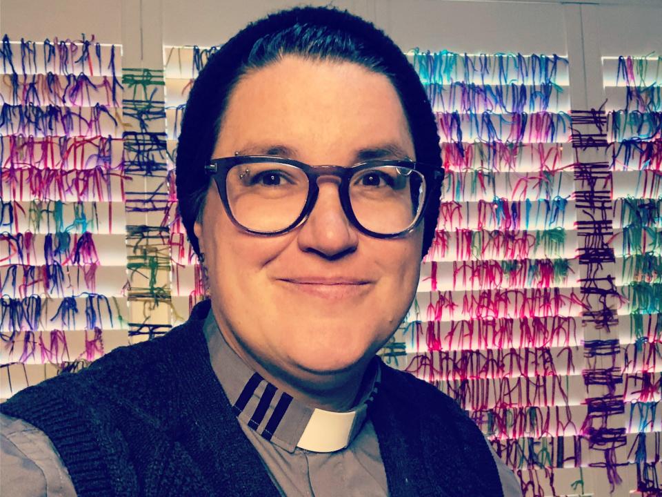 Rev. Megan Rohrer wears glasses and a beanie and takes a selfie in front of a rainbow-colored wall.