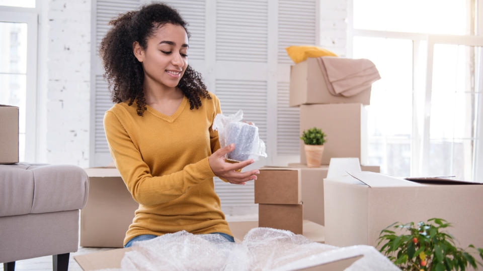 Safety measures. Beautiful curly-haired girl wrapping a tea cup securely before putting it into a box and smiling charmingly