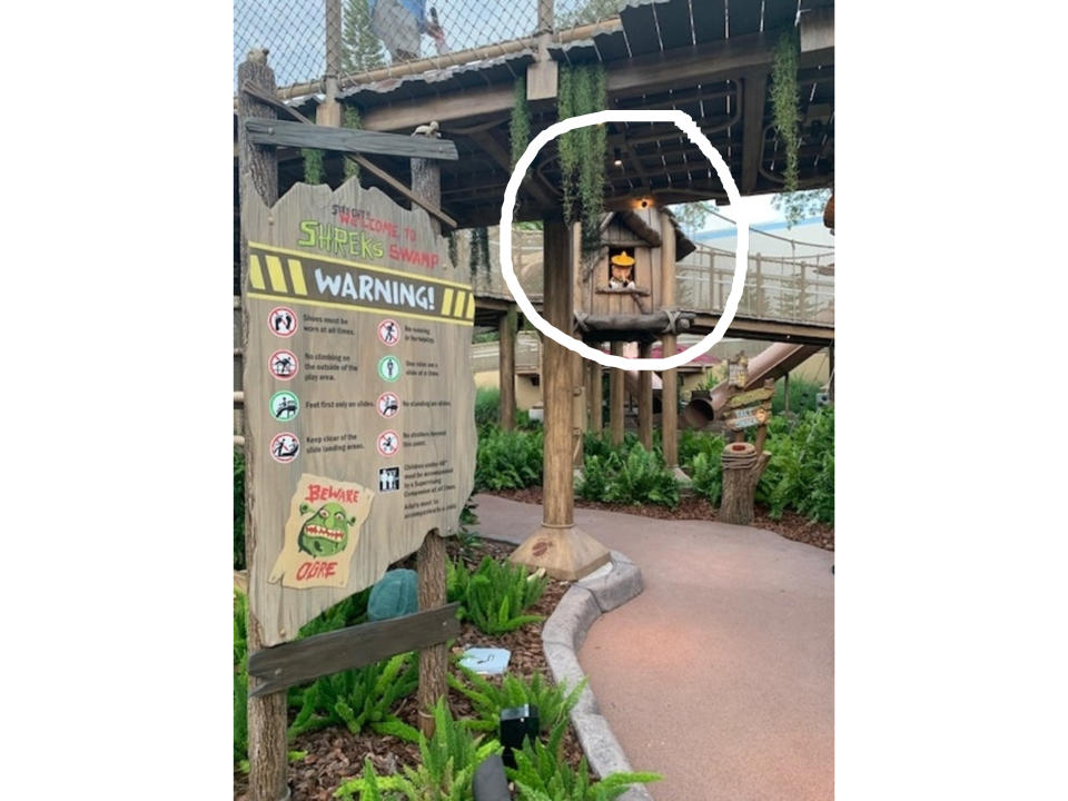 Wooden sign at Shrek's Swamp warns against various actions using illustrations. Behind it are wooden walkways and greenery, part of an amusement park setting