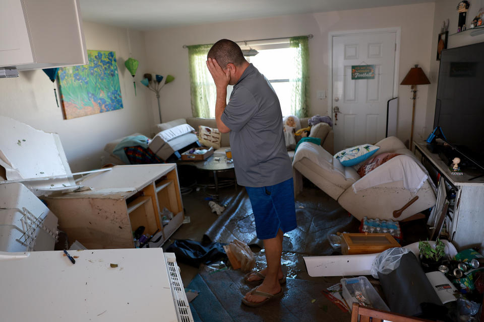 A man wearing a gray shirt, blue shorts, flip flops stands in a cluttered, destroyed living room and holds his head in his hands and cries