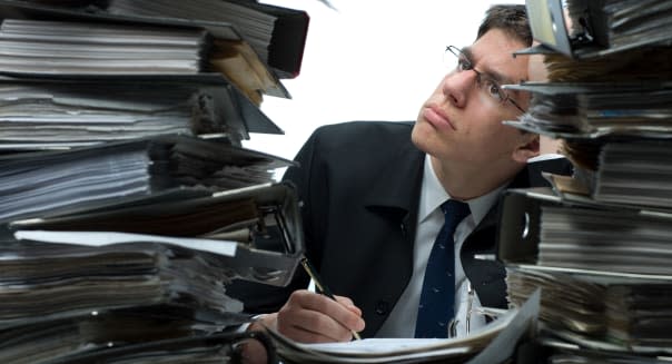 A confused man, holding a pen, sitting amidst piles of binders