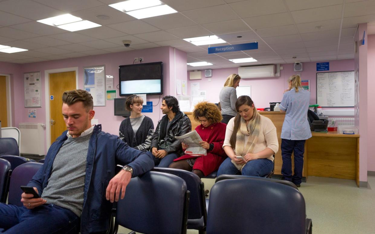 A mixed group of people can be seen waiting in a health clinic to be seen by the doctor. It is a typical British waiting room