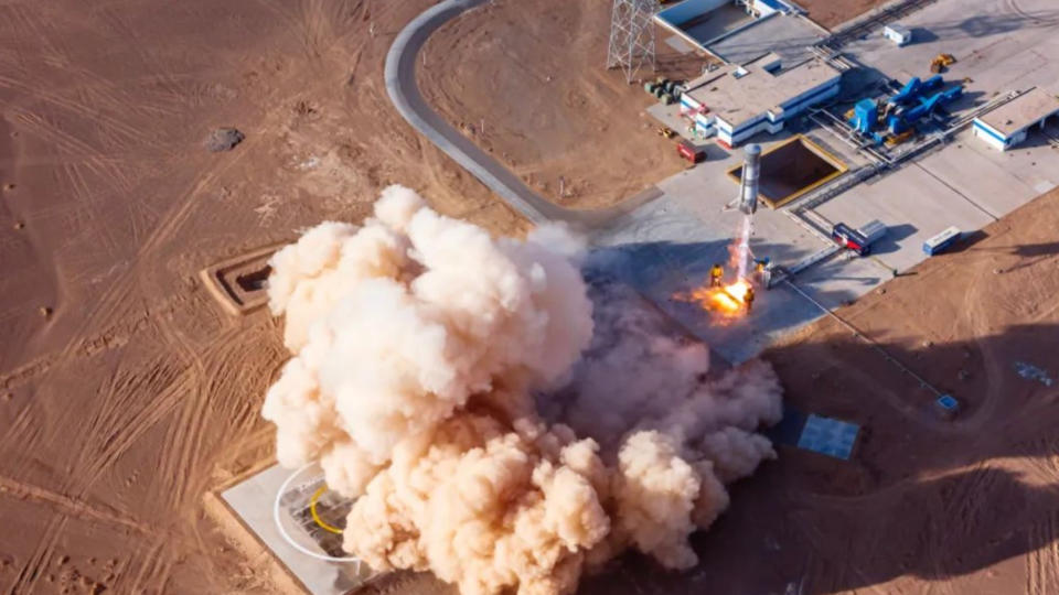 a small silver rocket launches from a gray pad in the middle of a desert landscape, raising a cloud of tan dust.