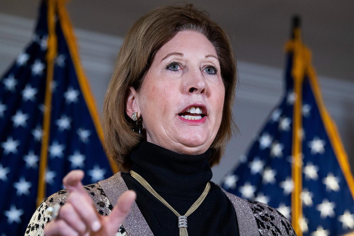 Powell speaking to the press with U.S. flags behind her wears a leopard print sweater vest, a black turtleneck sweater, and a necklace. 