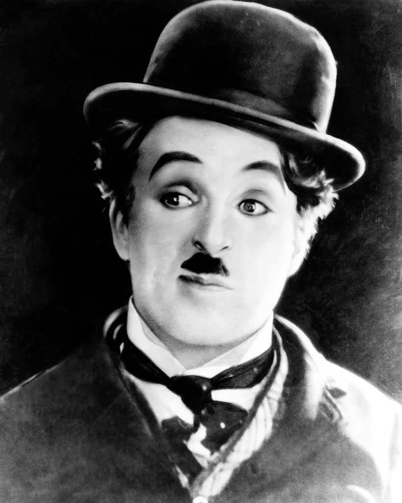 Charlie Chaplin, in his iconic Tramp costume, featuring a bowler hat, mustache, and bow tie, poses with a curious expression