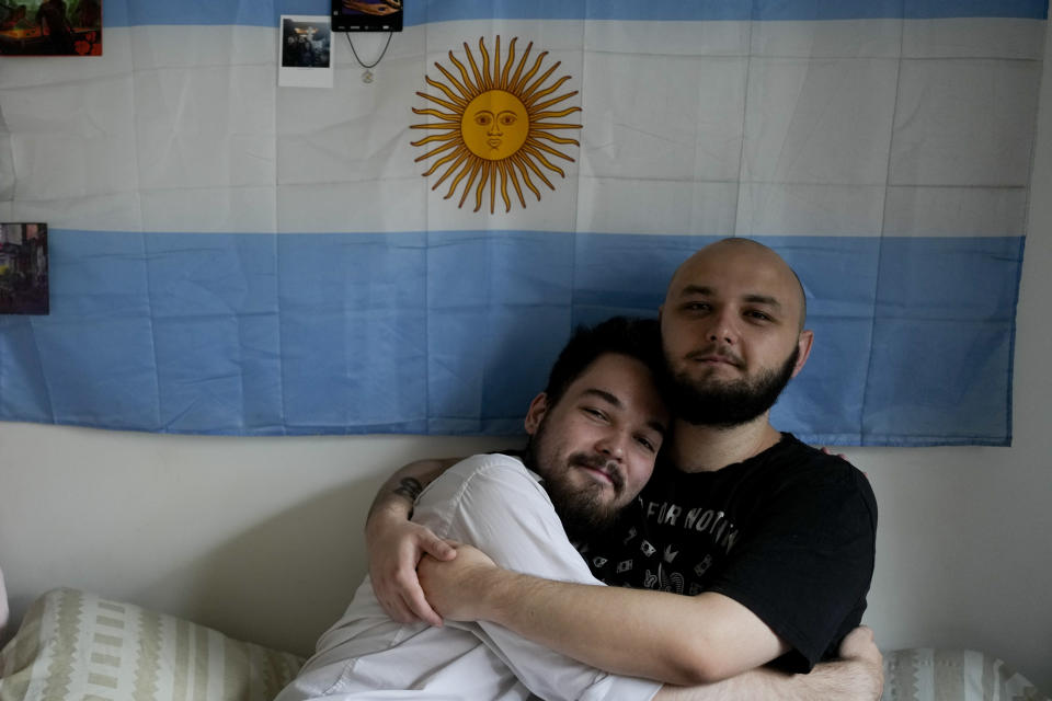 Russian nationals Dmitri Yarin, left, and his partner Nikolai Shushpan, embrace backdropped by an Argentine national flag as they pose for a picture at their home in Buenos Aires, Argentina, Monday, Feb. 13, 2023. Shuspan is enjoying living openly as a gay man for the first time. Back home, there was always “tension” and the feeling “that something could happen, said Shushpan. (AP Photo/Natacha Pisarenko)