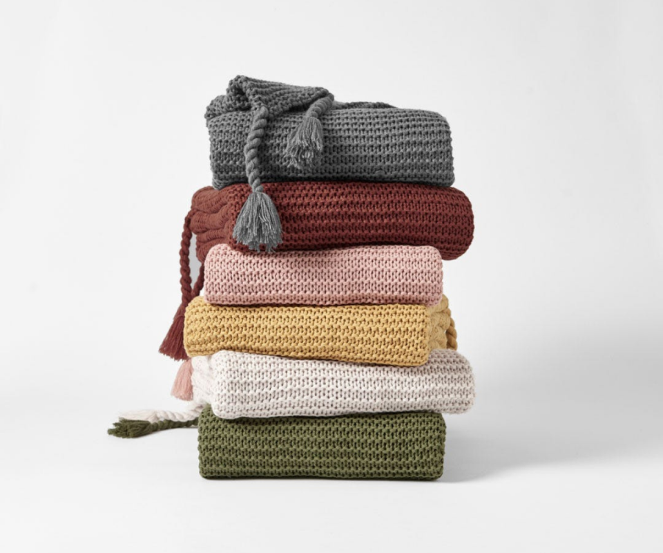 Six knitted chunky throw blankets in olive, cream, mustard, pale pink, maroon and grey are stacked ontop of each other on a white background.