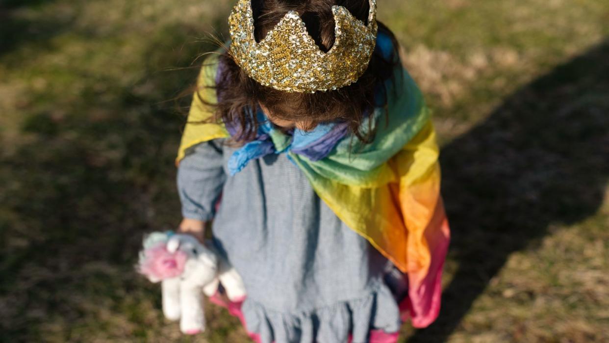 three year old girl with crown and rainbow cape on carrying unicorn toy