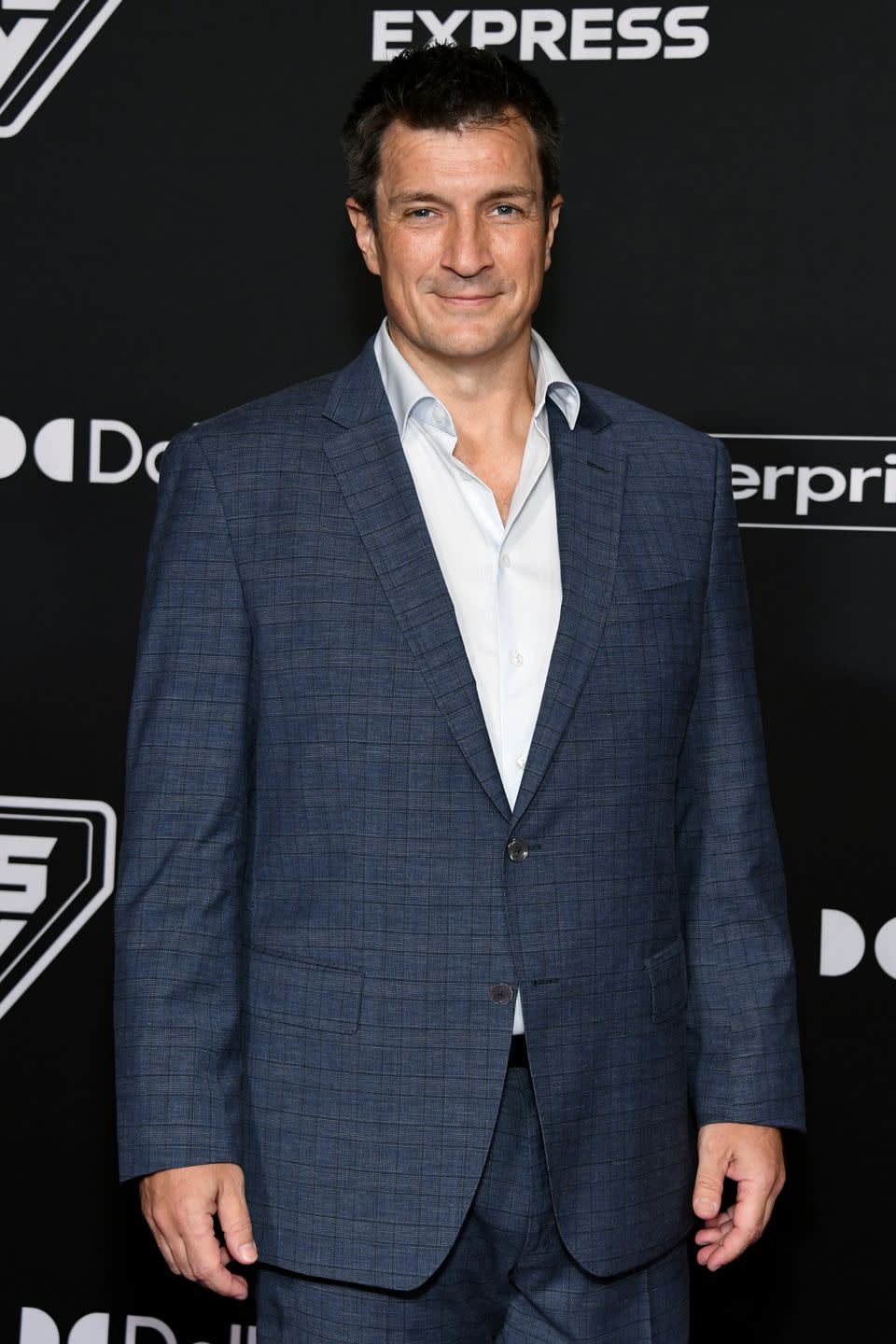 nathan fillion wearing a white collared shirt and a navy blue suit, standing in front of a black background at the world premiere of guardians of the galaxy volume 3 in april 2023