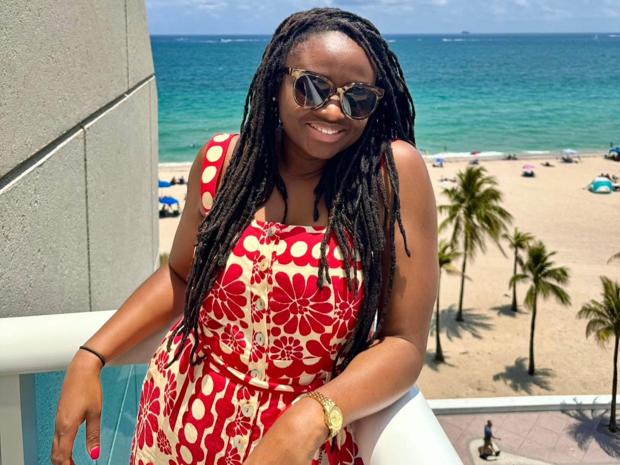 Mariette wears sunglasses and a red-and-white-patterned romper on a balcony overlooking the beach.