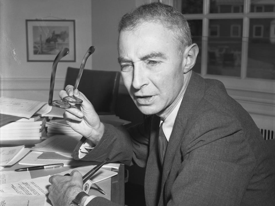 J.Robert Oppenheimer sits at a desk holding a pair of glasses.