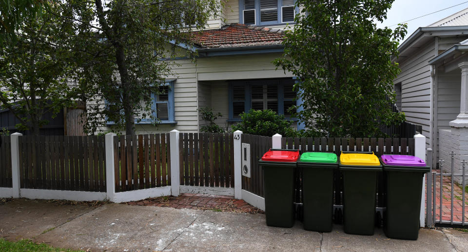 Four rubbish bins with red, green, yellow and purple lids outside a house.