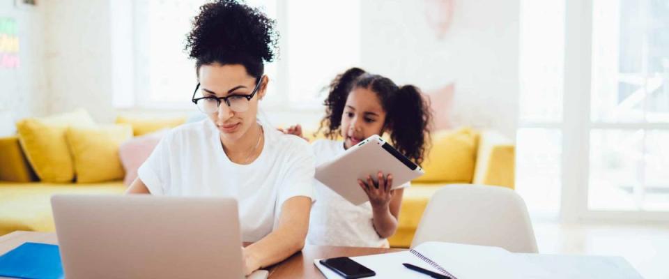 Little girl standing in bright room and showing tablet to mother while parent in glasses working in front of laptop.