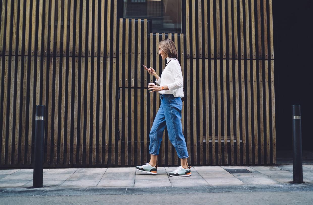 Side view of concentrated female pedestrian in stylish outfit text messaging on mobile phone while walking along pavement near contemporary building