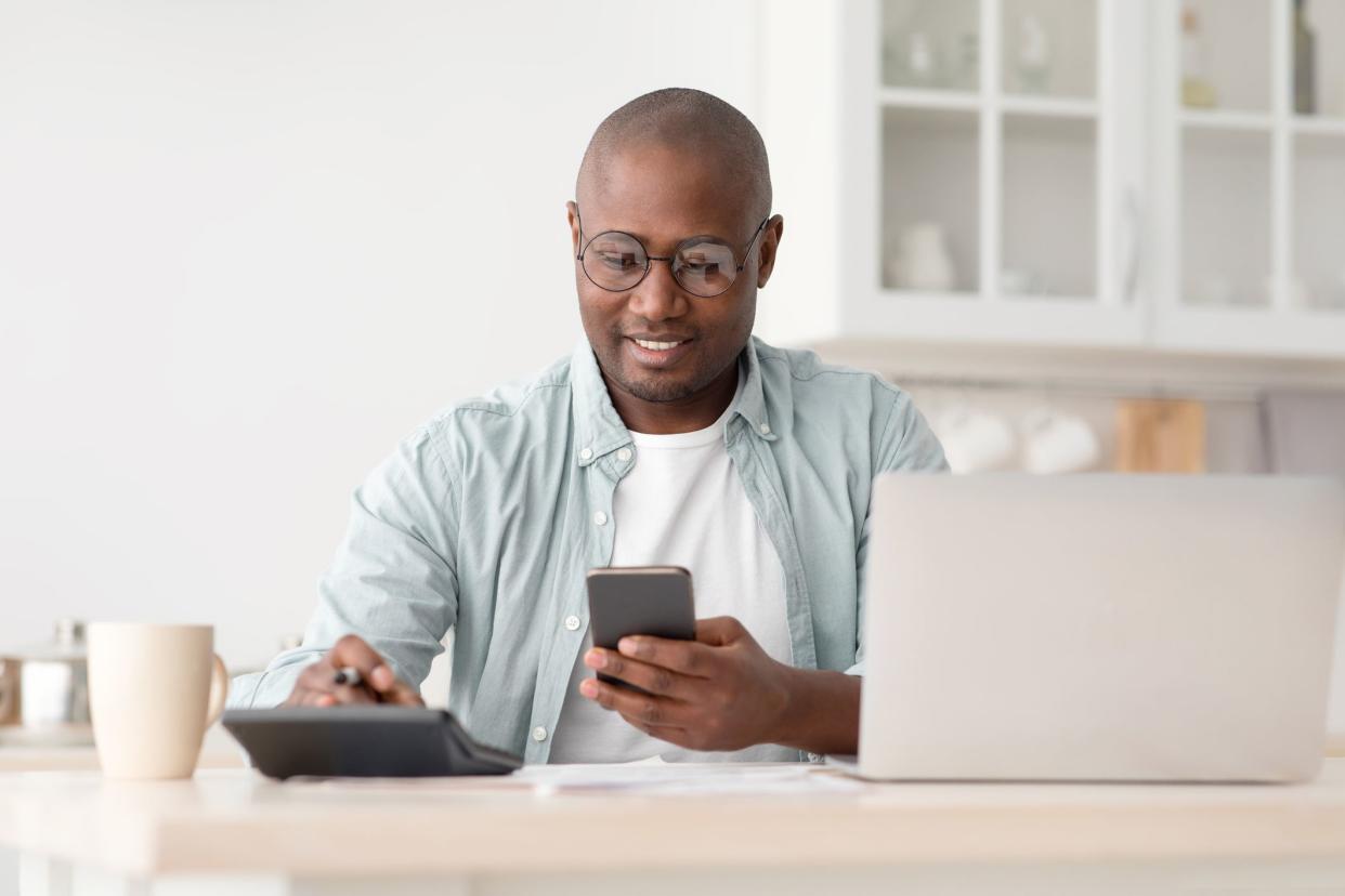 Savings and finances concept. Mature black man using calculator, phone and laptop computer, calculating taxes, sitting in kitchen at home, copy space