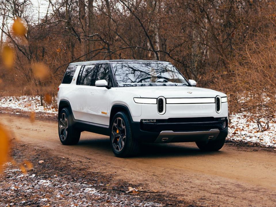 A white Rivian R1S parked on a dirt road with bare trees in the background.