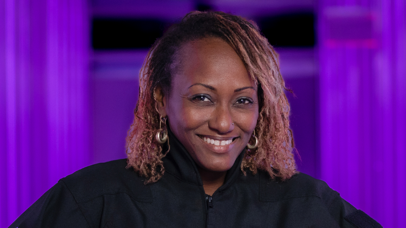 A black woman smiles for a portrait photo against a purple background.