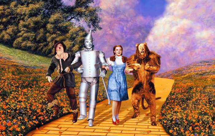 Scarecrow, Tin Man, Dorothy, and Cowardly Lion walk on a yellow brick road in a scenic landscape with flowers and trees, inspired by "The Wizard of Oz"