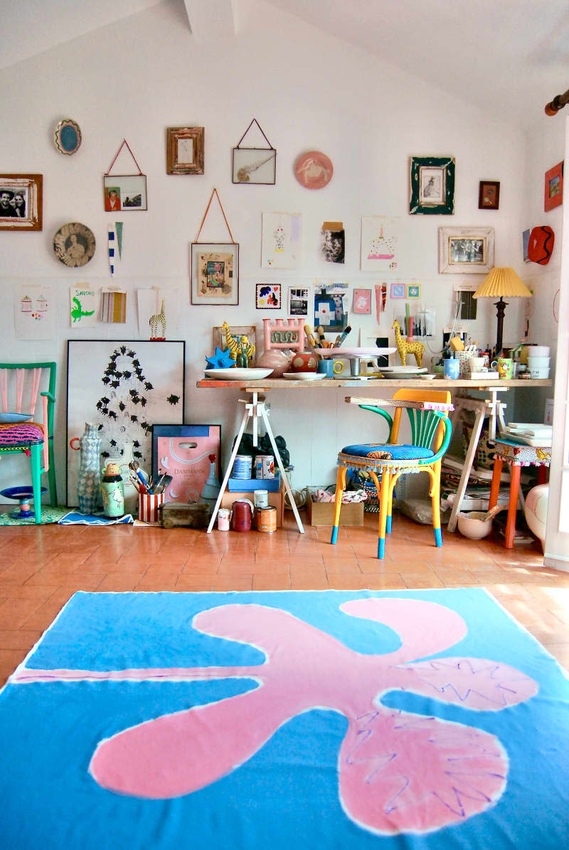 Art supplies on desk in white room with gallery walls and blue and pink artwork on floor.