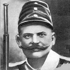 Béla Kiss, wearing a Hungarian military hat and holding a gun, and sporting a very wide mustache.