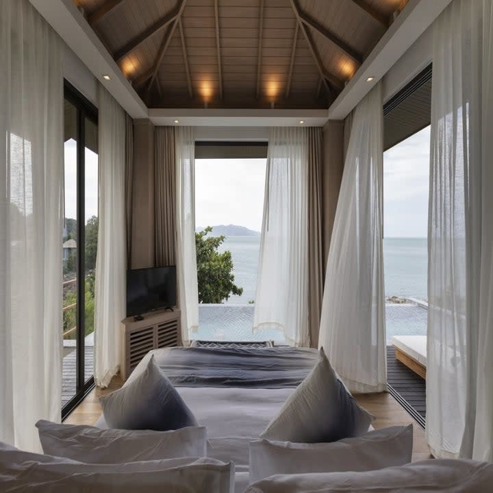 Hotel room with bed and floor to ceiling windows on all sides allowing a panoramic view of the ocean, bedroom leads out to private infinity pool
