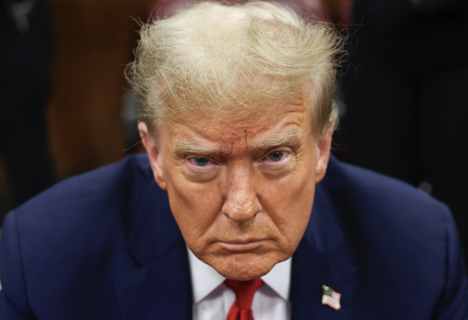 Donald Trump looks directly at the camera with a serious expression. He is wearing a blue suit and red tie