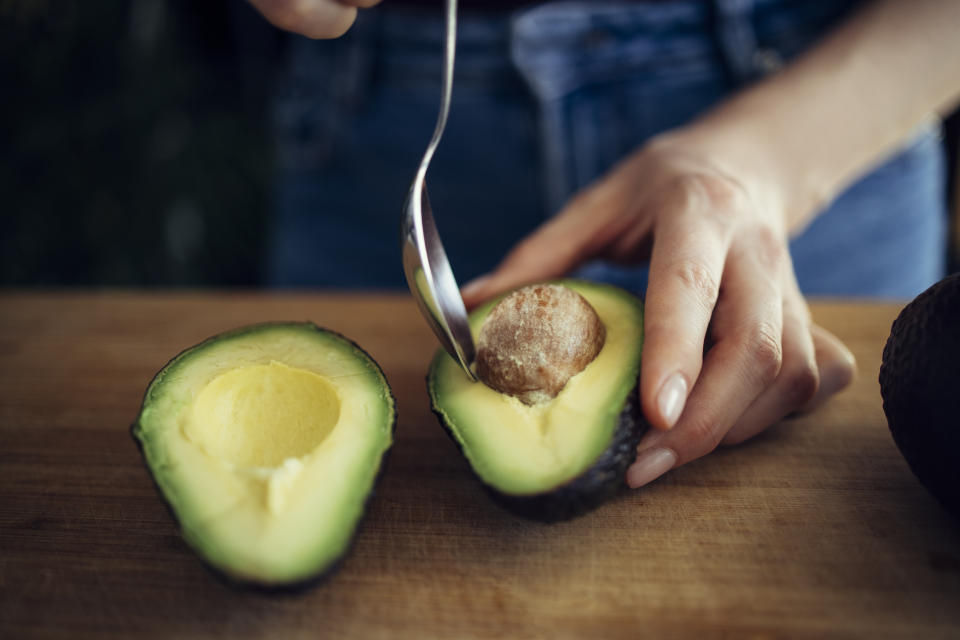 Woman's hands removing seed from avocado with spoon on wooden cutting board