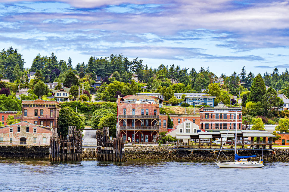 Coastal town with waterfront buildings, wooden piers, and a sailboat on the water. Trees and houses are in the background under a partly cloudy sky