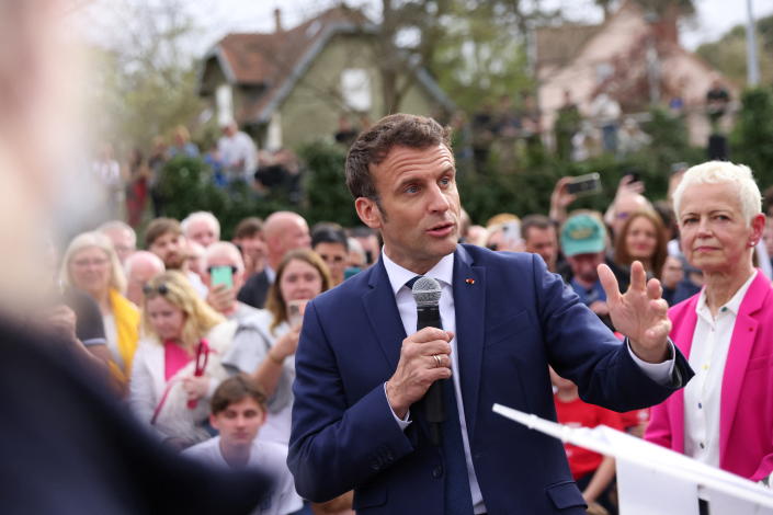 President Emmanuel Macron in full swing holds a microphone at a rally while a pensive Brigitte Klinkert listens on the sidelines.