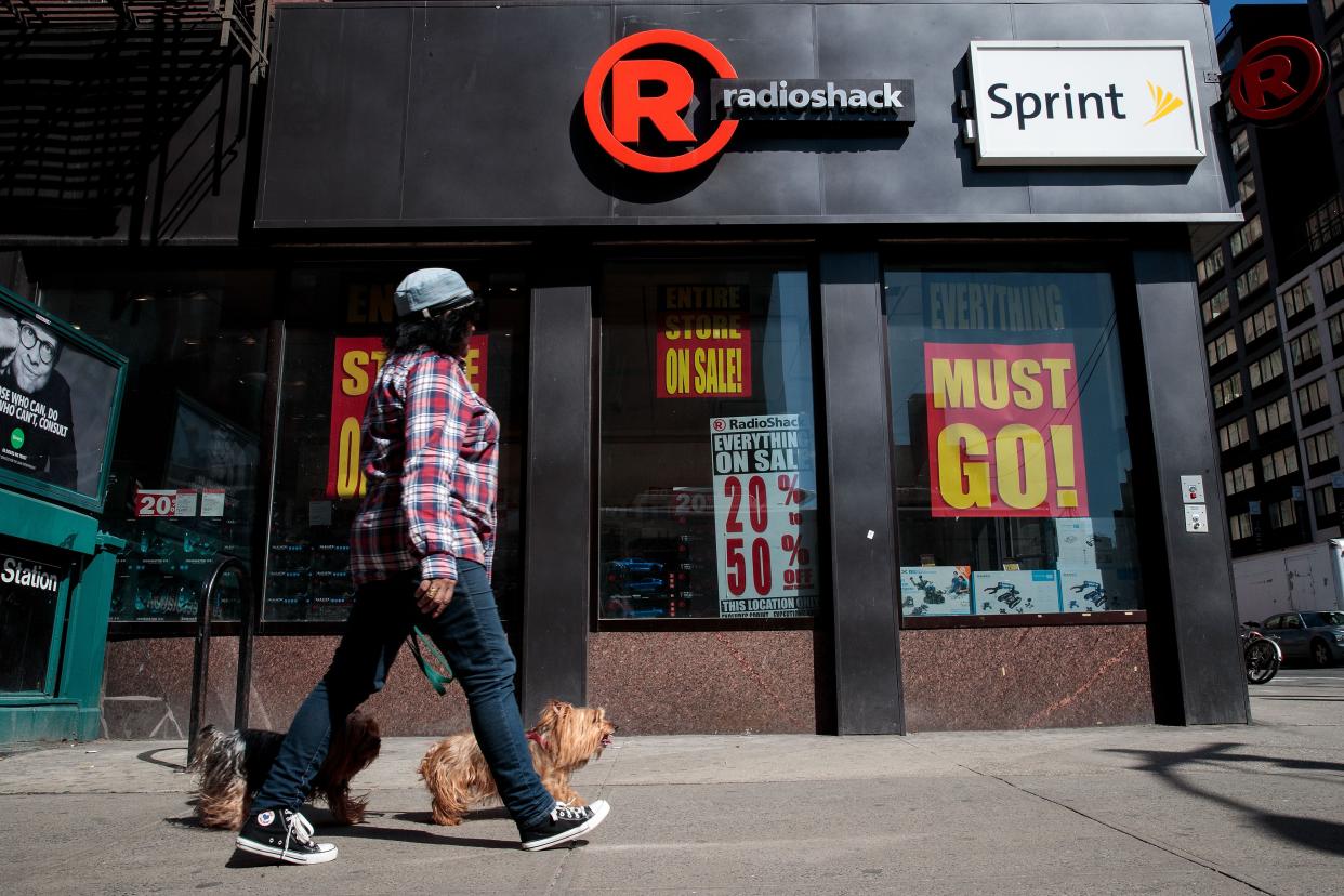 Front exterior of RadioShack in Chelsea neighborhood of Manhattan, New York City with many store closing sale signs in the windows, a woman is walking her two dogs along the sidewalk in the foreground, on a sunny day