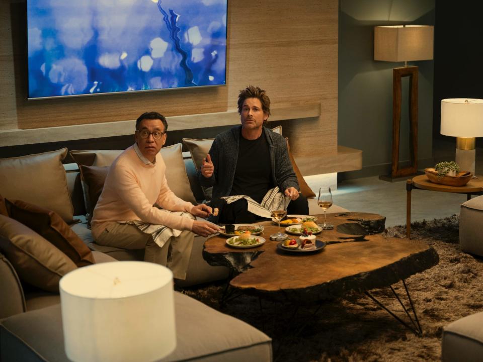 Fred Armisen and Rob Lowe sit in a nicely furnished living room with plates of food in front of them on "Unstable"