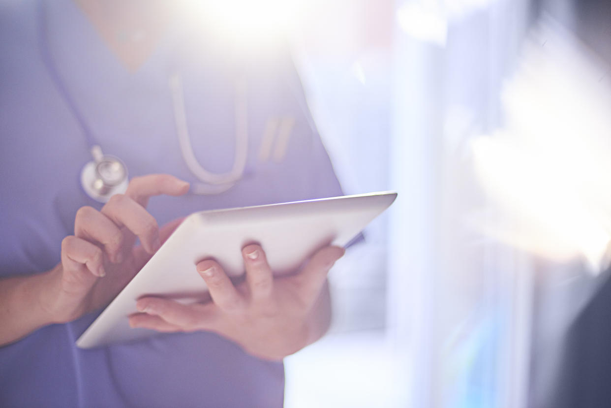 Female surgeon stood with her arms folded in hospital or surgery. She is wearing scrubs and has her stethoscope around her neck, the sun is bursting over her shoulder. She is looking at her patients records on her digital tablet and sending emails.